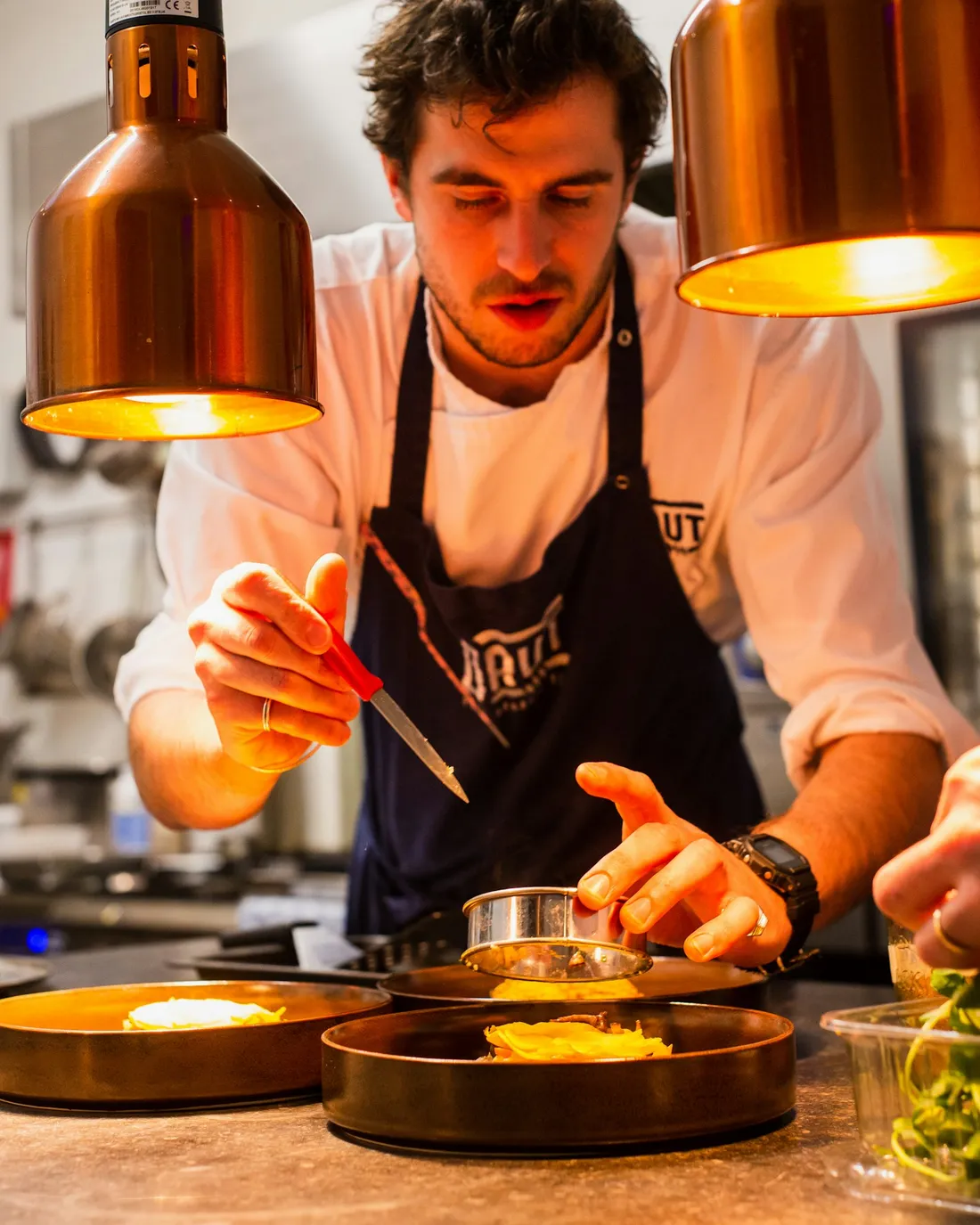 Cocinero emplatando con pinzas bajo lámparas cálidas
